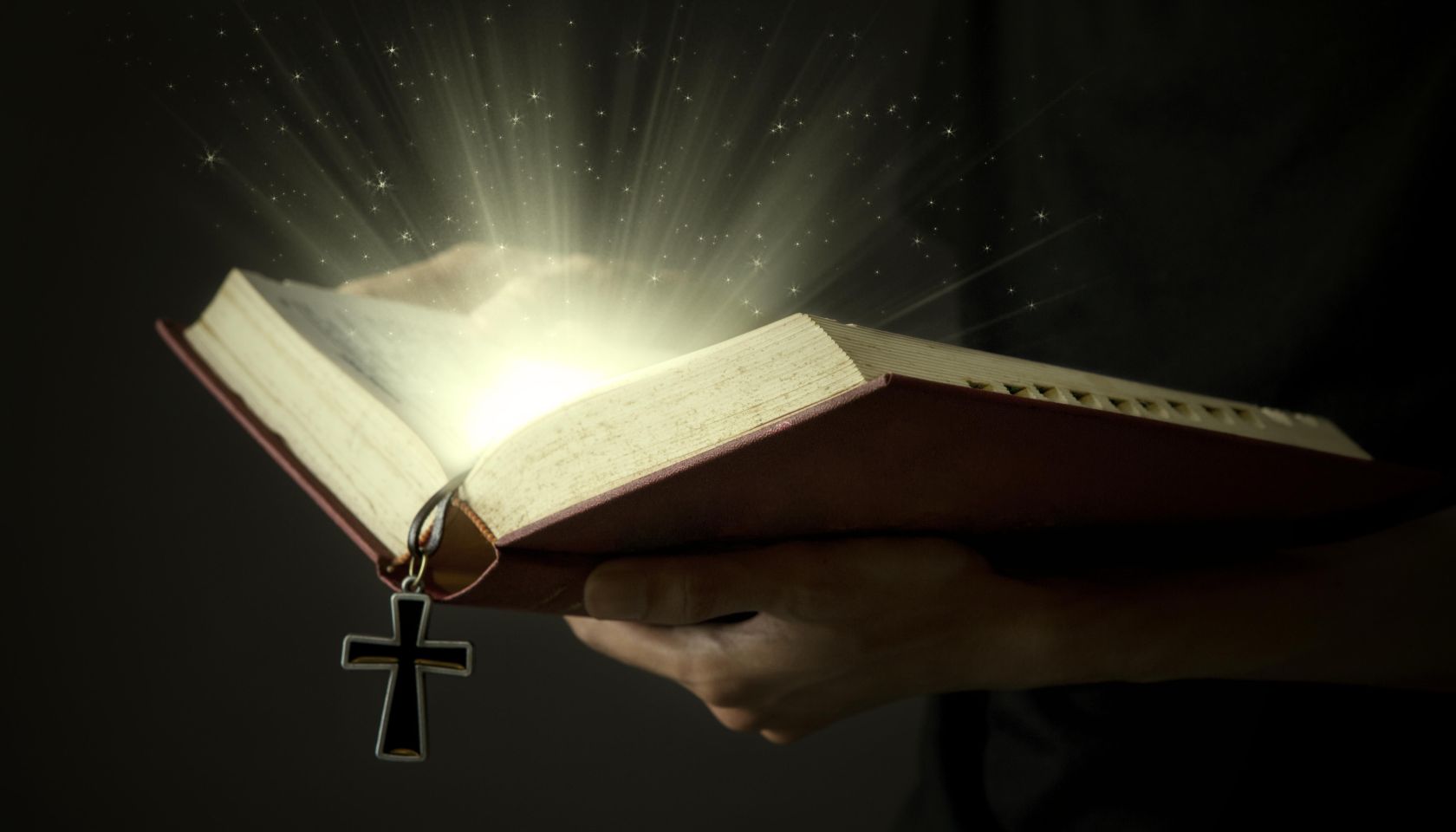 Hands of man holding holy bible and wooden rosary with magical rays