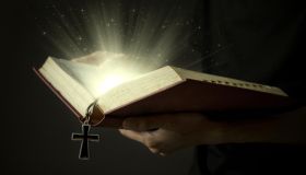 Hands of man holding holy bible and wooden rosary with magical rays