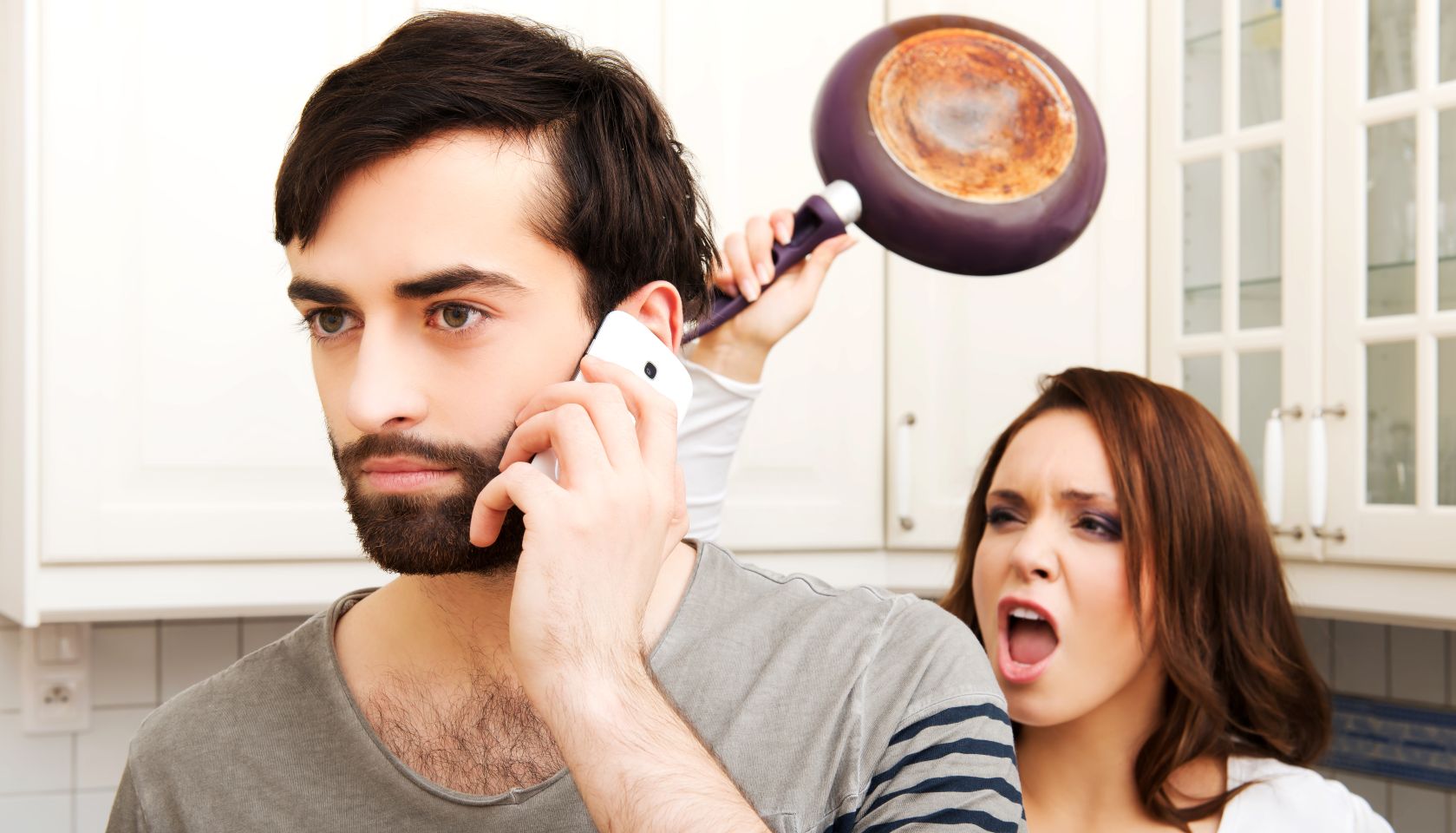 Young Woman Attacking Boyfriend With Cooking Pan In Kitchen At Home