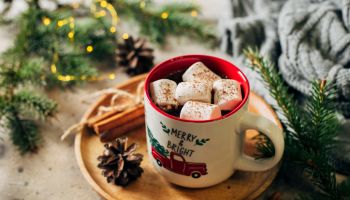 Hot chocolate with marshmallows and candy cane in holiday mug. Fir tree branch, cozy gray sweater on background. Cozy seasonal Christmas holidays concept