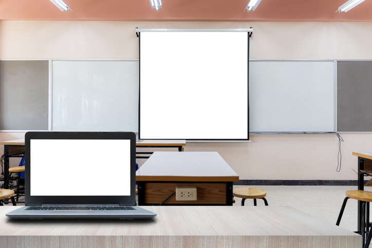 Laptop on table foreground. Projector screen canvas in modern conference room with big windows. Side view. Empty classroom or presentation room interior with desks