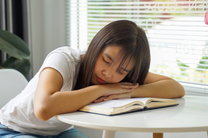 Asian woman sits reading a book and falls asleep on her hands clasped together on the book.