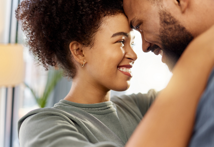 Happy mixed race couple hugging while relaxing at home. Carefree hispanic husband and wife smiling while staring into each other’s eyes lovingly and hugging in the lounge at home