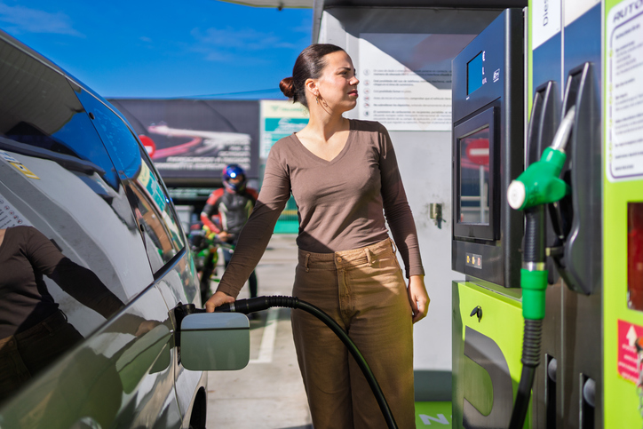 Woman contemplating rising gas prices and environmental impact while refueling car on sunny day. The intersection of transportation, energy, and the environment in everyday life