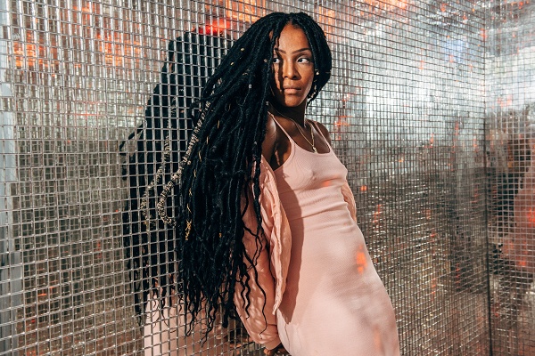 A woman with long dreadlocks wearing a pink dress poses in front of a wire mesh backdrop.