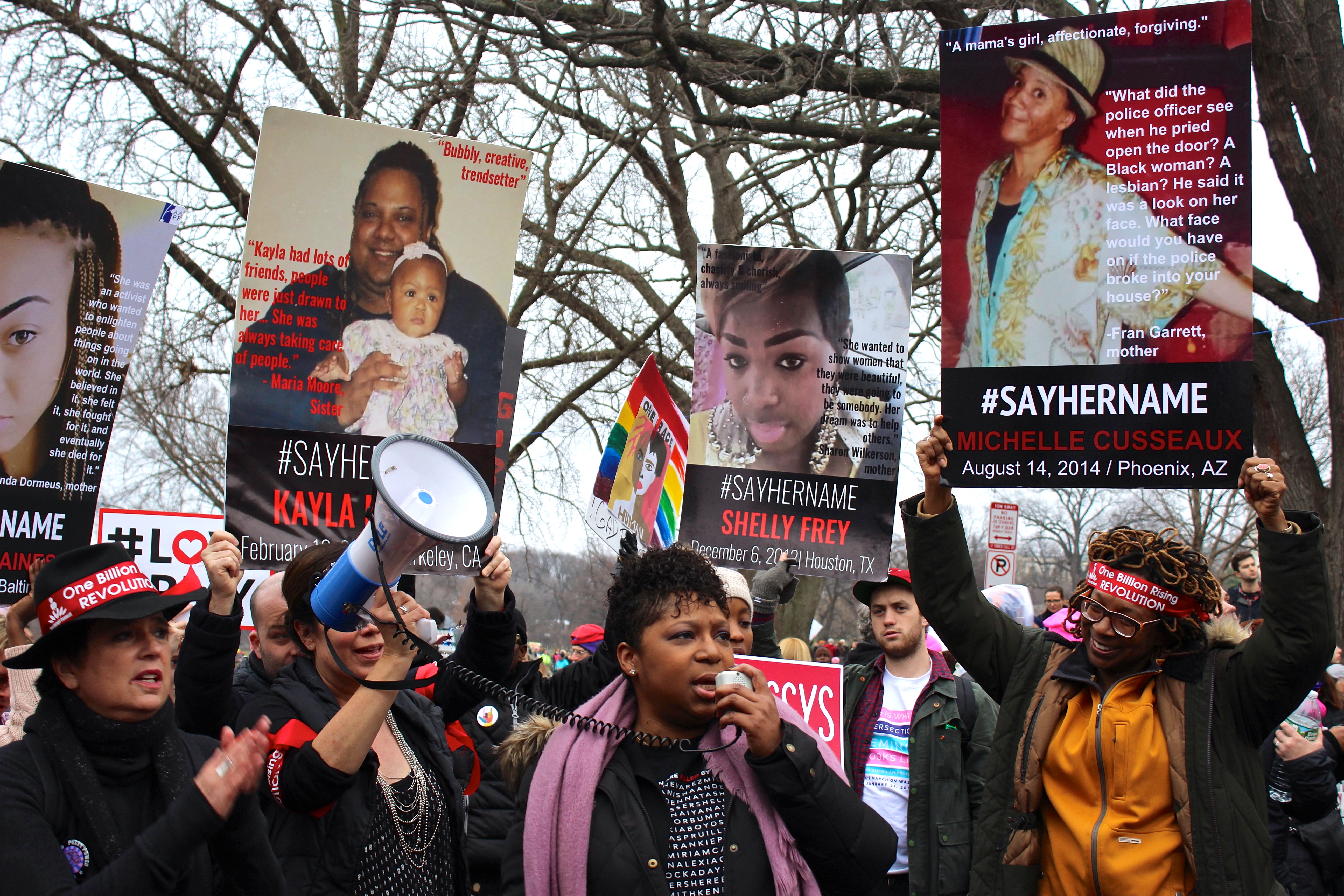 Protesters holding signs with names and slogans, including "Kayla", "Shelly Frey", and "#SAYHERNAME", in a public demonstration.