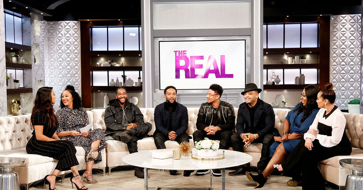 A group of seven people, including men and women, sitting on a couch in a television studio set with the text "THE REAL" displayed on a screen behind them.
