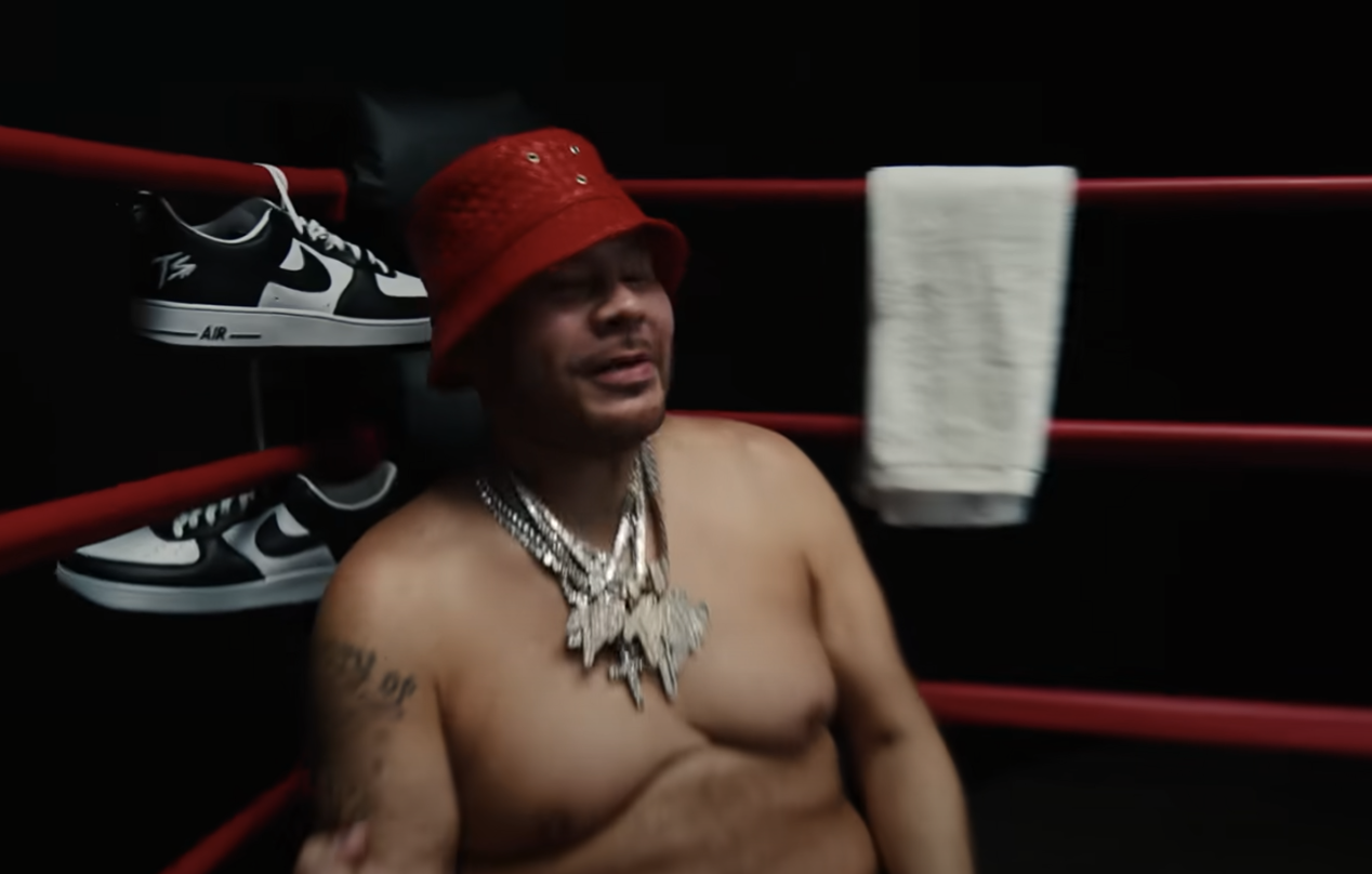 A person wearing a red hat and necklace standing in a boxing ring with Nike shoes visible.