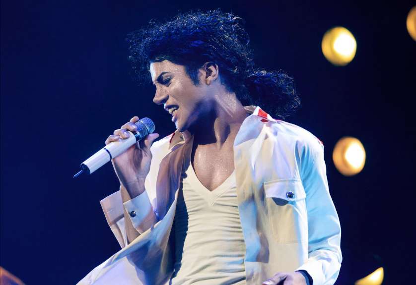 A performer with curly hair singing into a microphone on stage, wearing a white outfit.