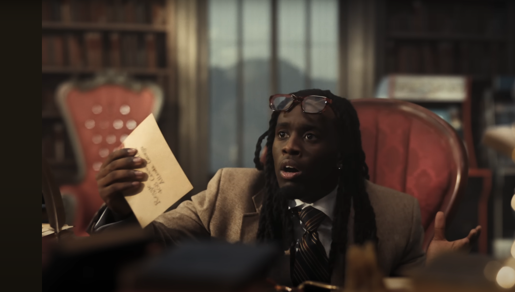 A person with dreadlocks wearing glasses and a suit, holding an envelope, sitting in a chair in a room with bookshelves.