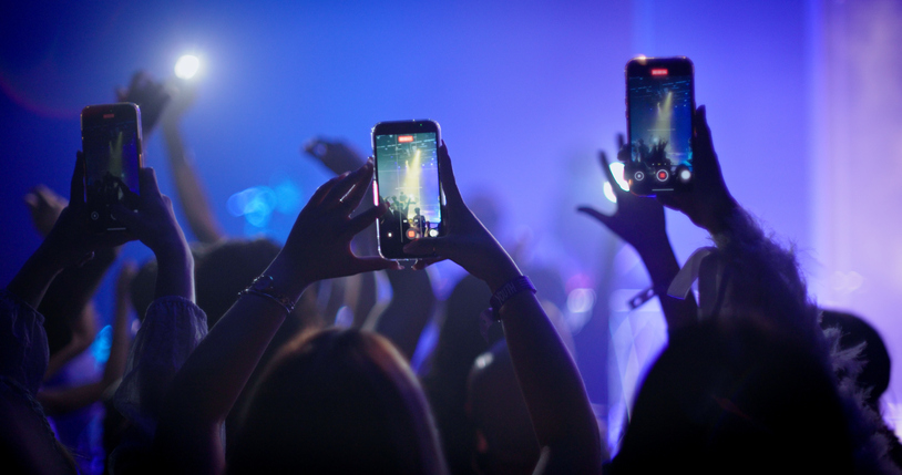 Silhouettes of music fans holding up mobile phones and smartphones to record video or take photos at a live concert.