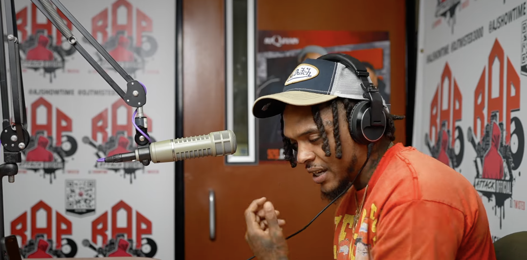 A person wearing a baseball cap and headphones speaking into a microphone in a recording studio with graffiti-style artwork on the walls.