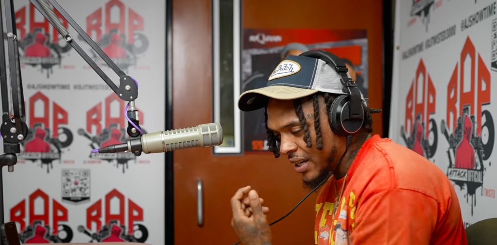 A person wearing a baseball cap and headphones speaking into a microphone in a recording studio with graffiti-style artwork on the walls.