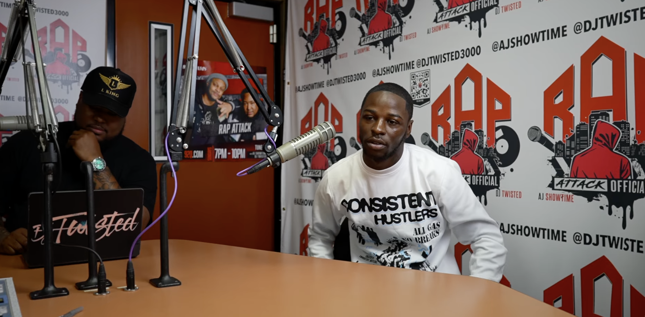 A man wearing a "Consistent Hustlers" t-shirt sitting at a microphone in a recording studio with "Rap Attack" and "DJ Twisted 3000" branding visible on the walls.