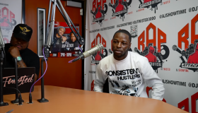 A man wearing a "Consistent Hustlers" t-shirt sitting at a microphone in a recording studio with "Rap Attack" and "DJ Twisted 3000" branding visible on the walls.