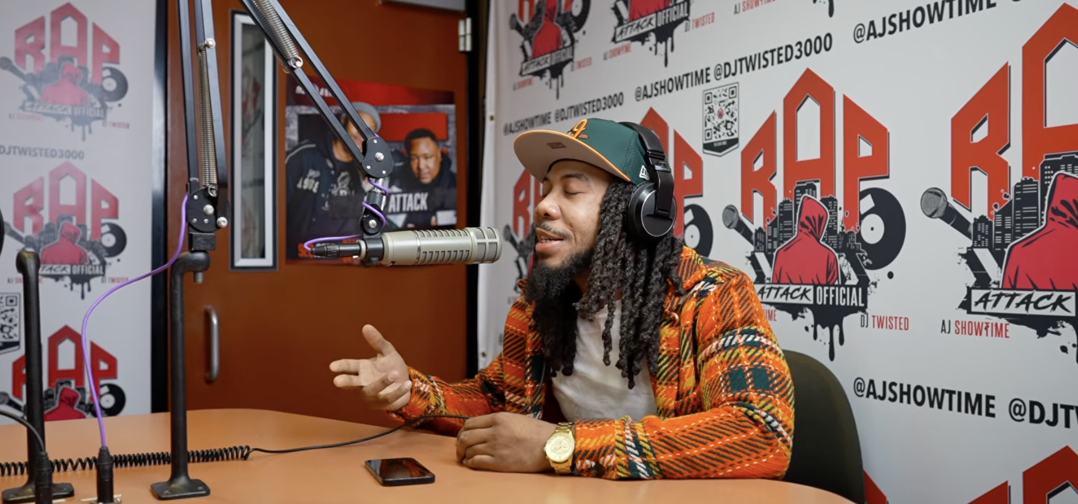 A person with dreadlocks wearing headphones and an orange and black patterned shirt, sitting at a microphone and speaking into it, in a studio setting with "RAP ATTACK" branding visible.