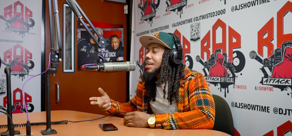 A person with dreadlocks wearing headphones and an orange and black patterned shirt, sitting at a microphone and speaking into it, in a studio setting with "RAP ATTACK" branding visible.