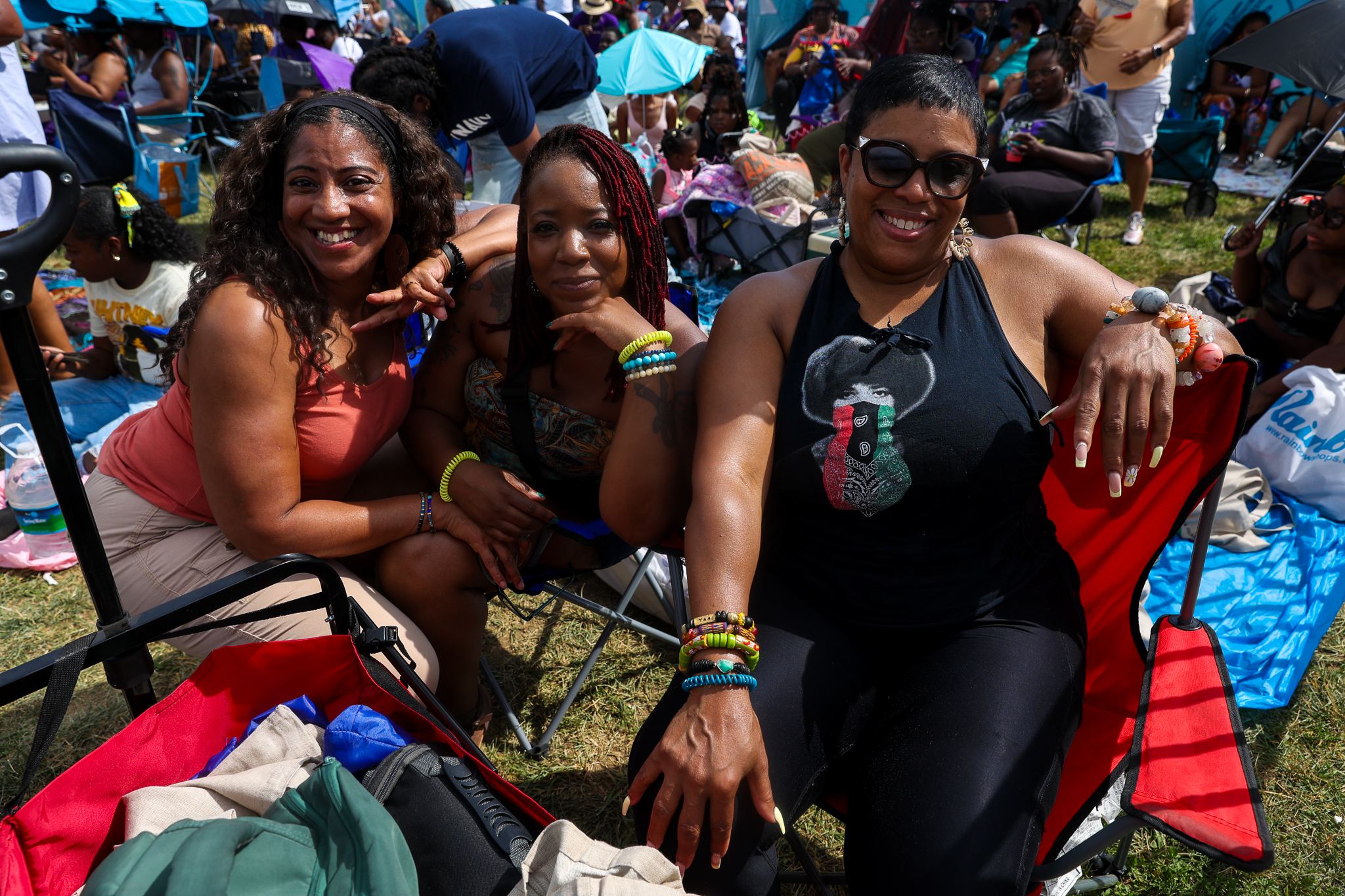 Three smiling women seated in lawn chairs at an outdoor event, wearing colorful clothing and accessories.