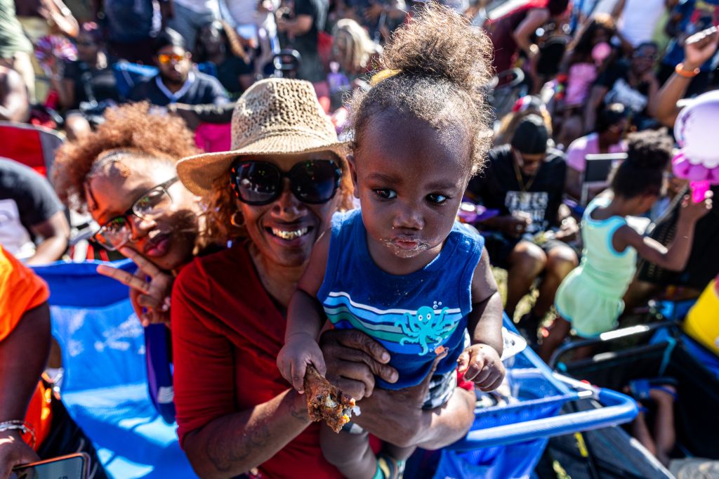 A smiling woman in a red shirt holding a young child in a blue shirt, surrounded by a crowd of people in a lively outdoor setting.