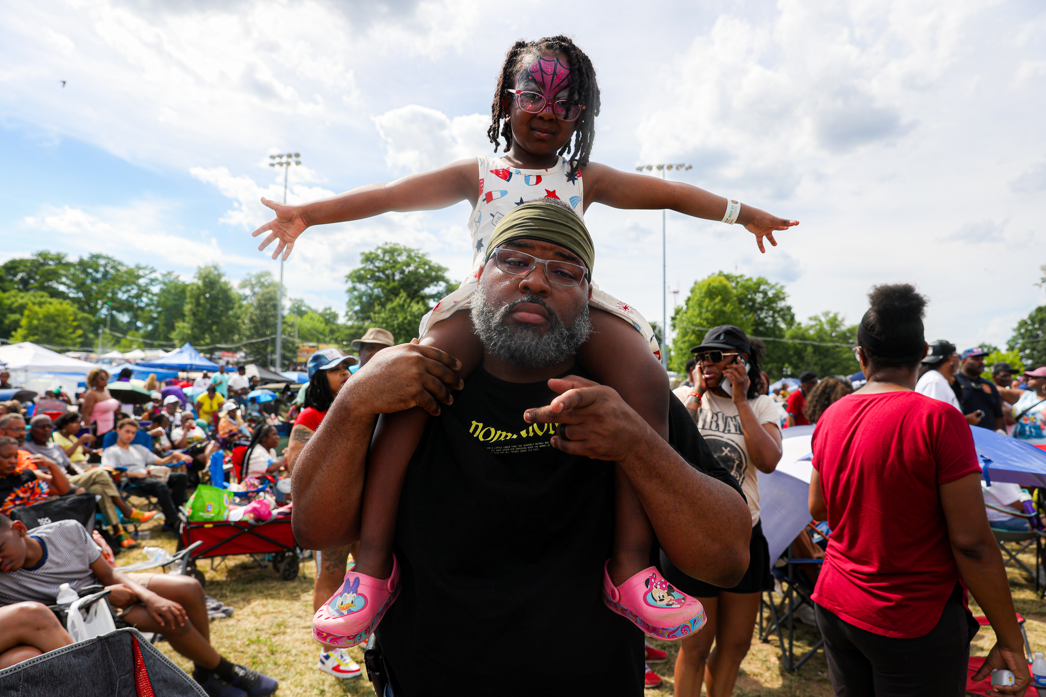 A man with a long beard carries a young child on his shoulders at a crowded outdoor event, with the child wearing a pink mask and colorful clothing.