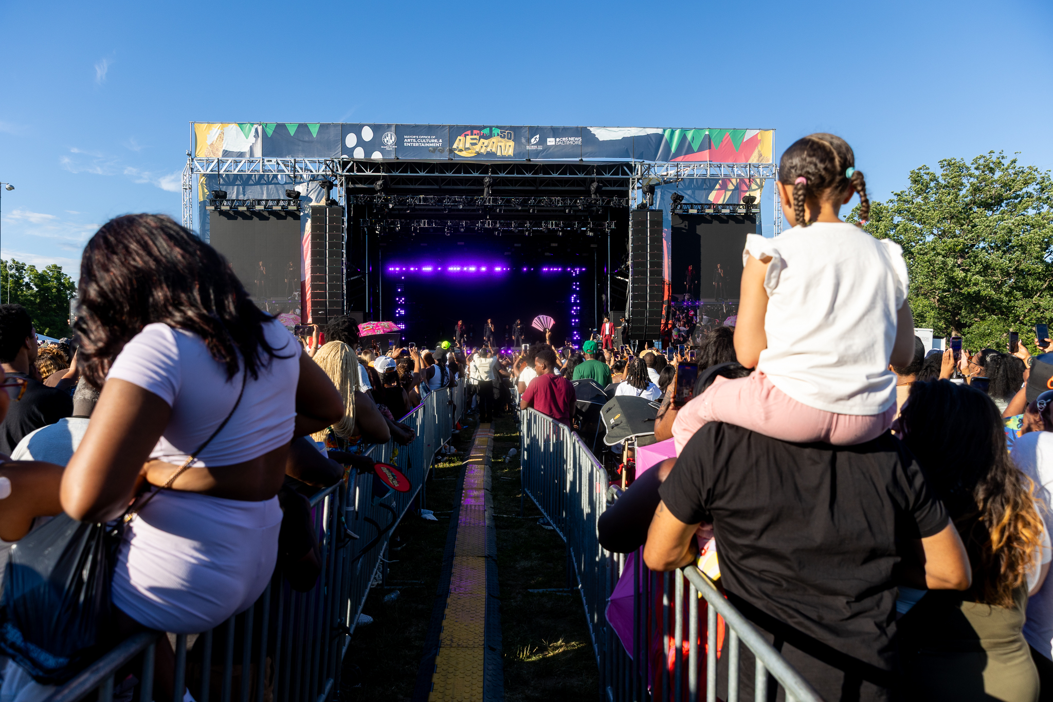 A large outdoor music festival stage with a colorful, patterned backdrop and a crowd of people in the foreground watching the performance.