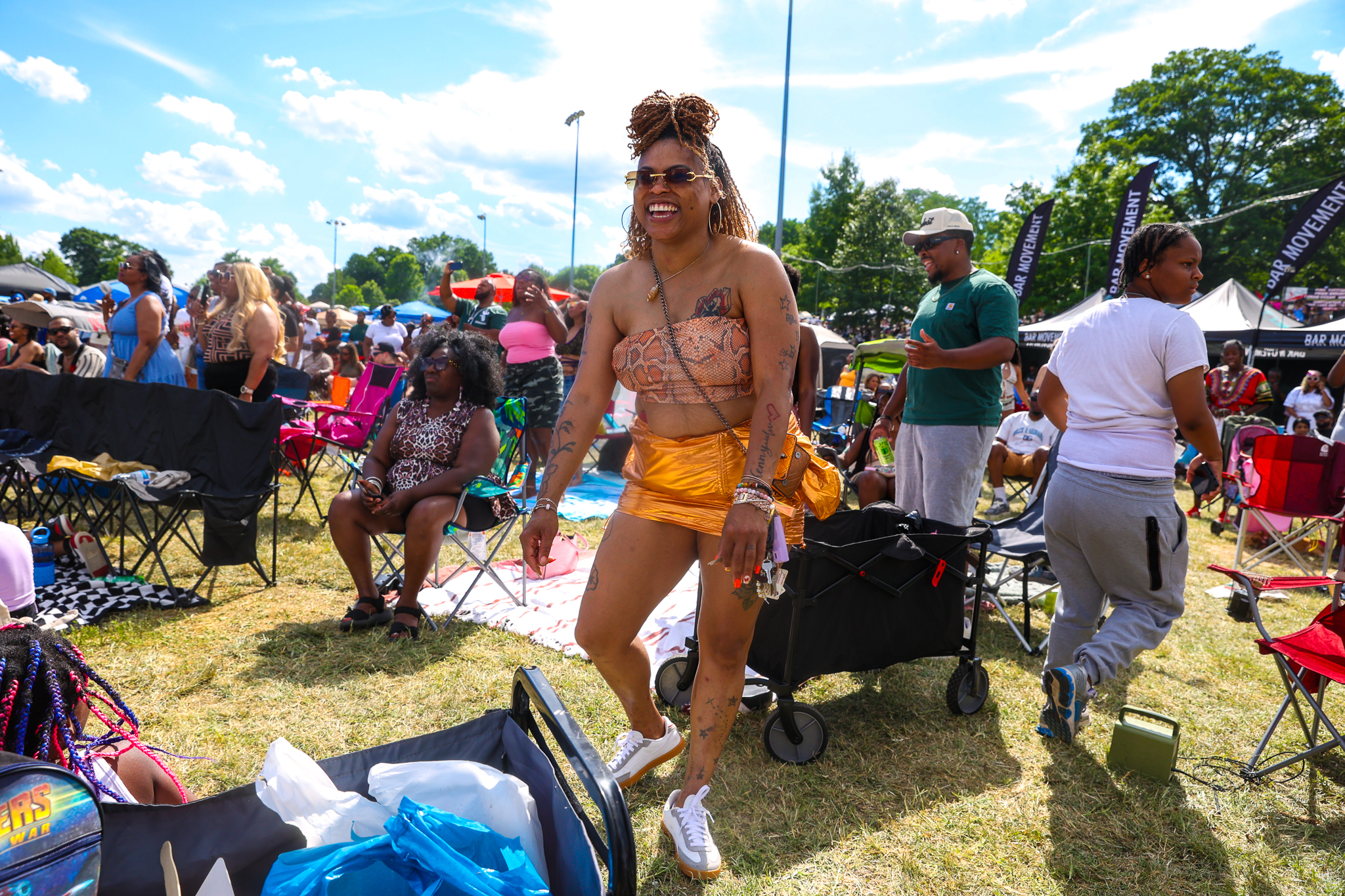 A smiling person wearing colorful clothing and accessories stands in a crowded outdoor festival setting with tents, chairs, and other people in the background.