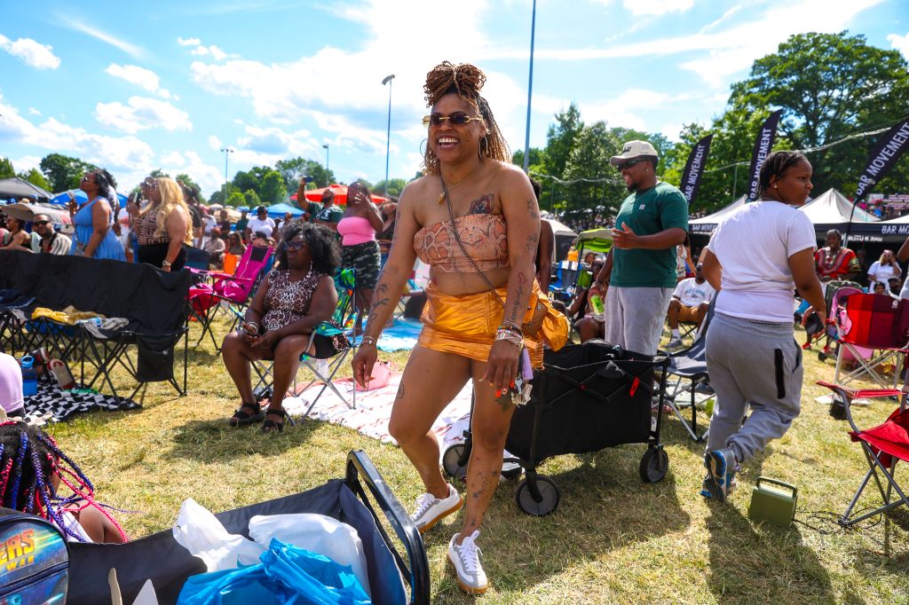 A smiling person wearing colorful clothing and accessories stands in a crowded outdoor festival setting with tents, chairs, and other people in the background.