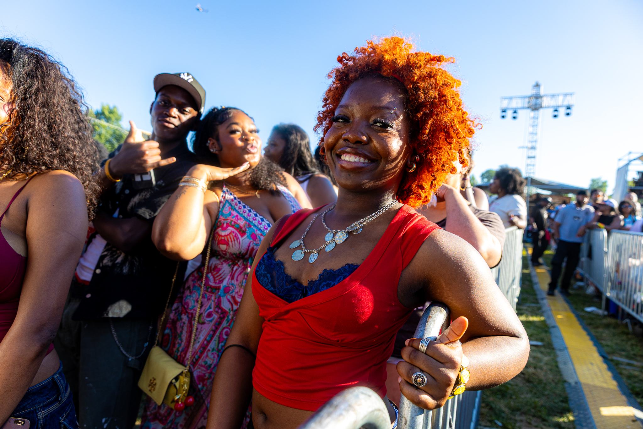 A group of smiling people at an outdoor event, with a woman in the foreground wearing a red top and bright orange curly hair.