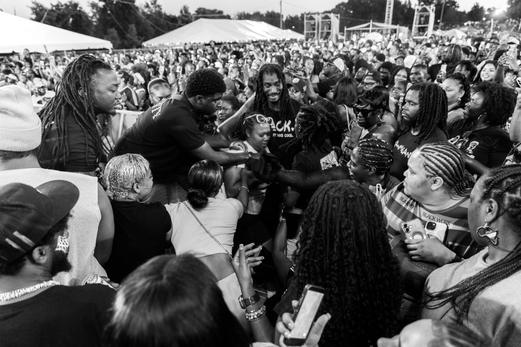 A large crowd of people, many with dreadlocks, gathered at an outdoor event or festival. Several people in the crowd are embracing and interacting with each other.