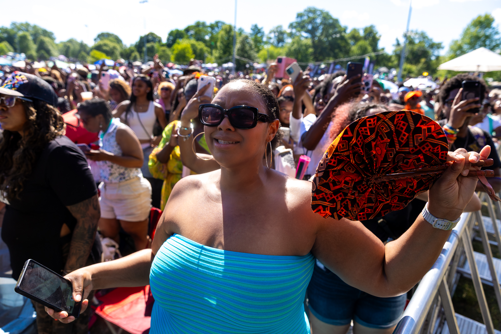 A large crowd of people at an outdoor event, with a woman in the foreground wearing sunglasses and holding a colorful patterned fan.