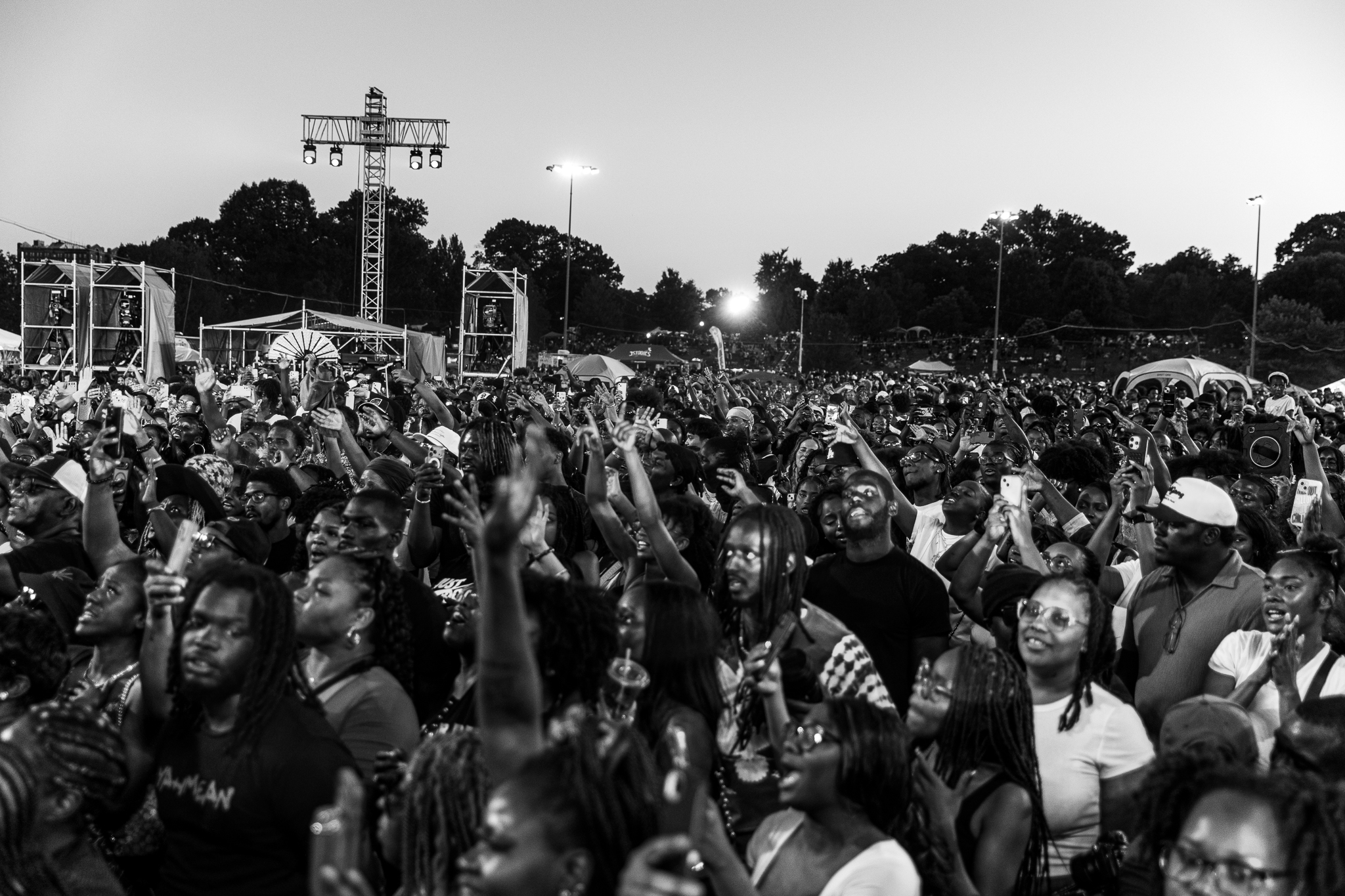 Large crowd of people with raised hands at a nighttime outdoor music festival or concert, with stage equipment and lighting visible in the background.
