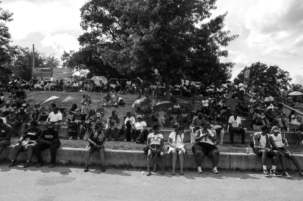 Crowded outdoor gathering with people sitting on steps and benches under trees on a cloudy day.