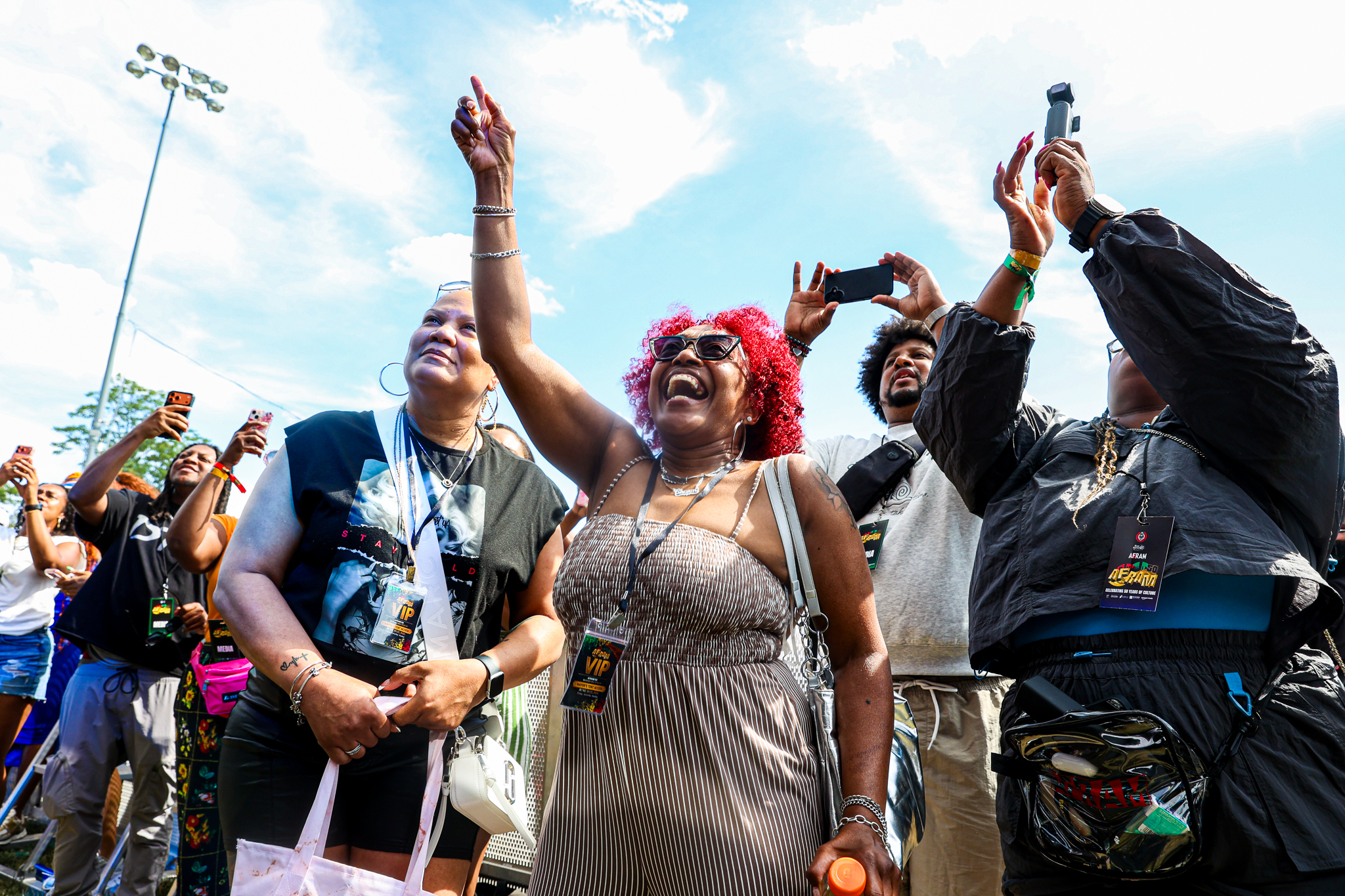Crowd of people at an outdoor event, some raising their arms and taking photos with smartphones. Several people wearing colorful clothing and accessories.