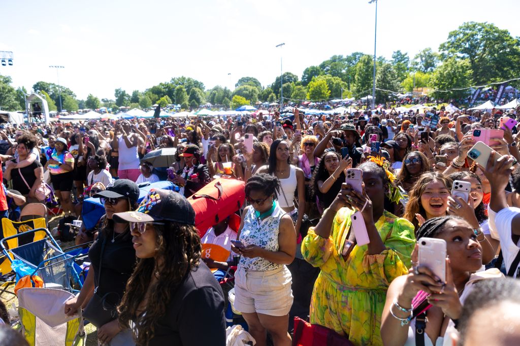 A large crowd of people gathered at an outdoor event, many holding up smartphones to take photos or videos. The crowd is diverse in age and attire, with some wearing colorful or festive clothing.