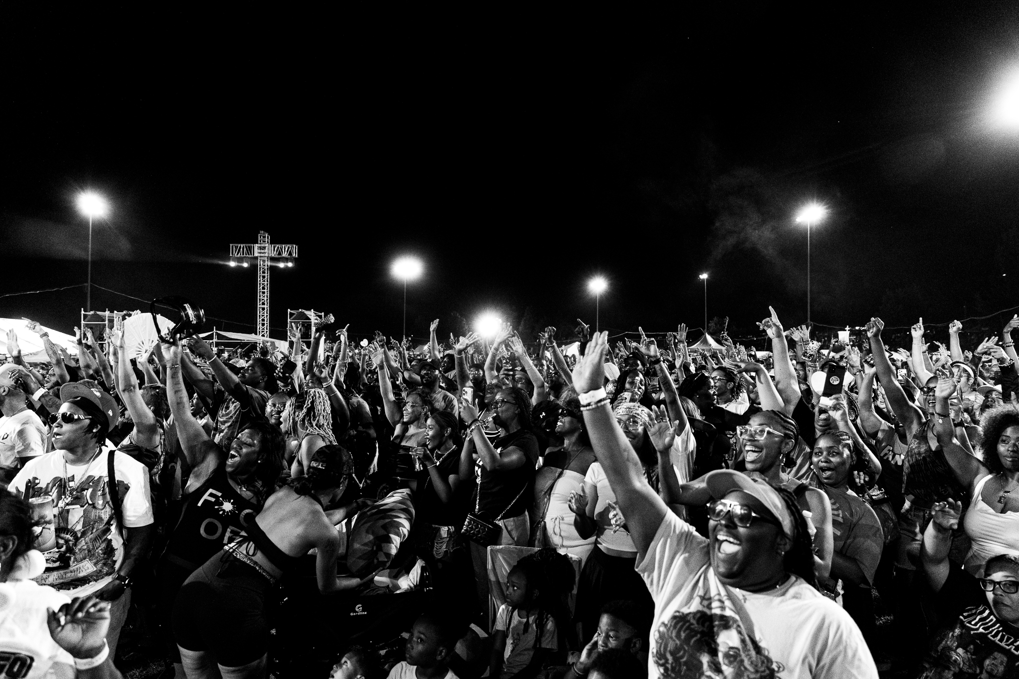 Nighttime crowd at a large outdoor event, with people raising their arms and cheering enthusiastically. The event appears to be a concert or festival based on the stage and lighting equipment visible.