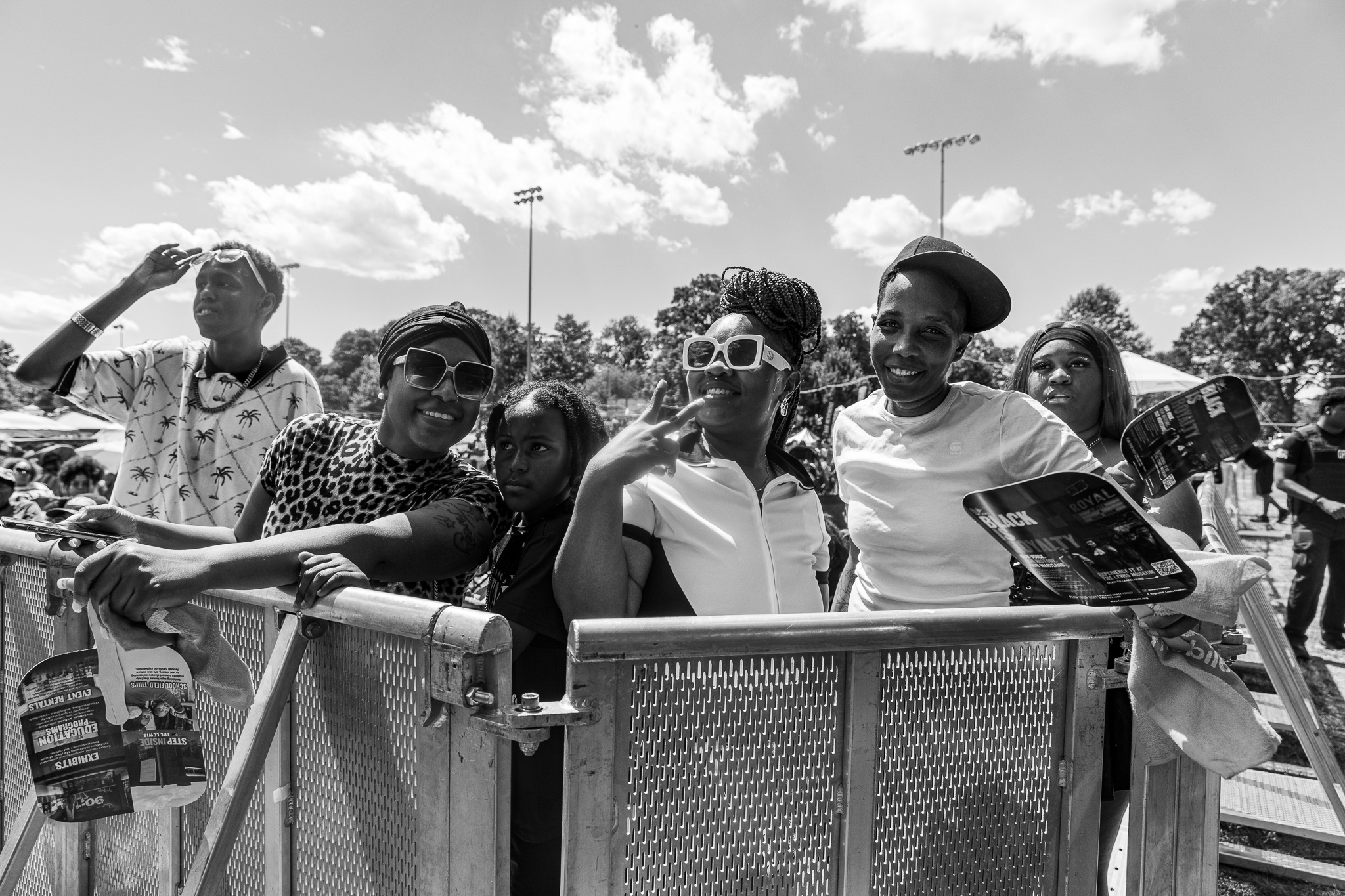 A group of smiling people, some wearing sunglasses, standing together behind a metal barrier at an outdoor event.