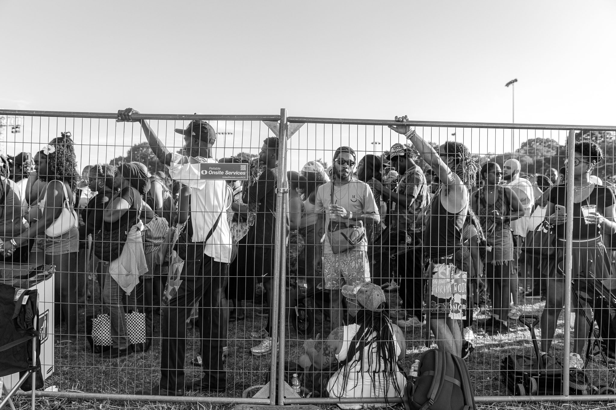 A crowd of people standing behind a metal fence, some holding signs or bags. The fence has "Onsite Services" printed on it.