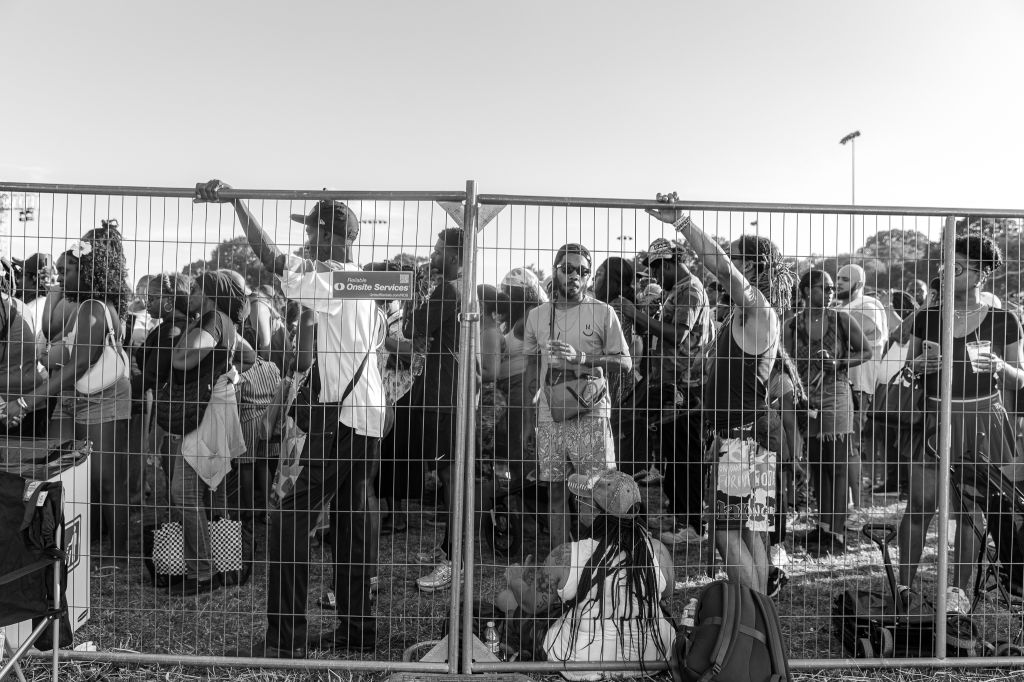 A crowd of people standing behind a metal fence, some holding signs or bags. The fence has "Onsite Services" printed on it.