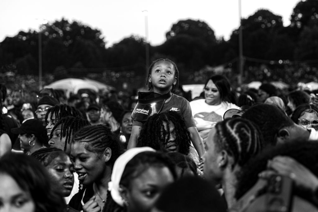 A crowd of people, mostly young Black individuals, gathered in an outdoor setting. A young girl wearing a Nike sweatshirt stands out in the foreground.