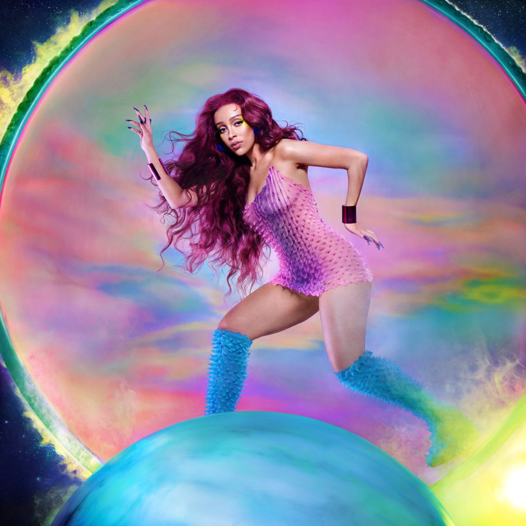 A person with long, wavy purple hair wearing a pink polka dot bodysuit and blue furry boots, posing in a colorful, psychedelic background.