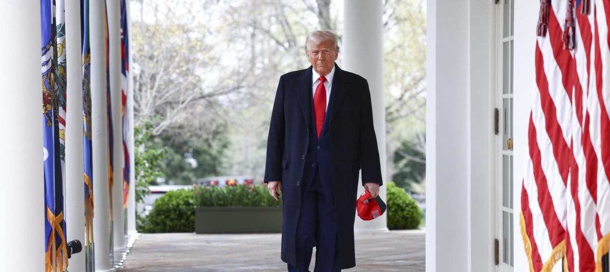 A man wearing a navy blue coat and red tie walking through the White House colonnade, with American flags and greenery visible.