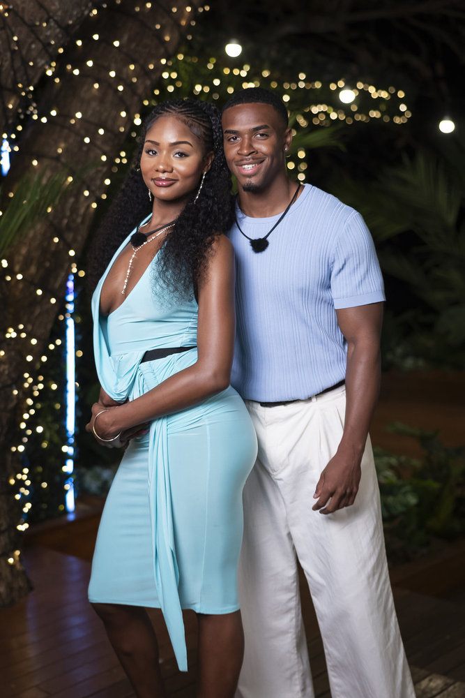 Two people, a woman and a man, standing together outdoors at night with holiday lights in the background.