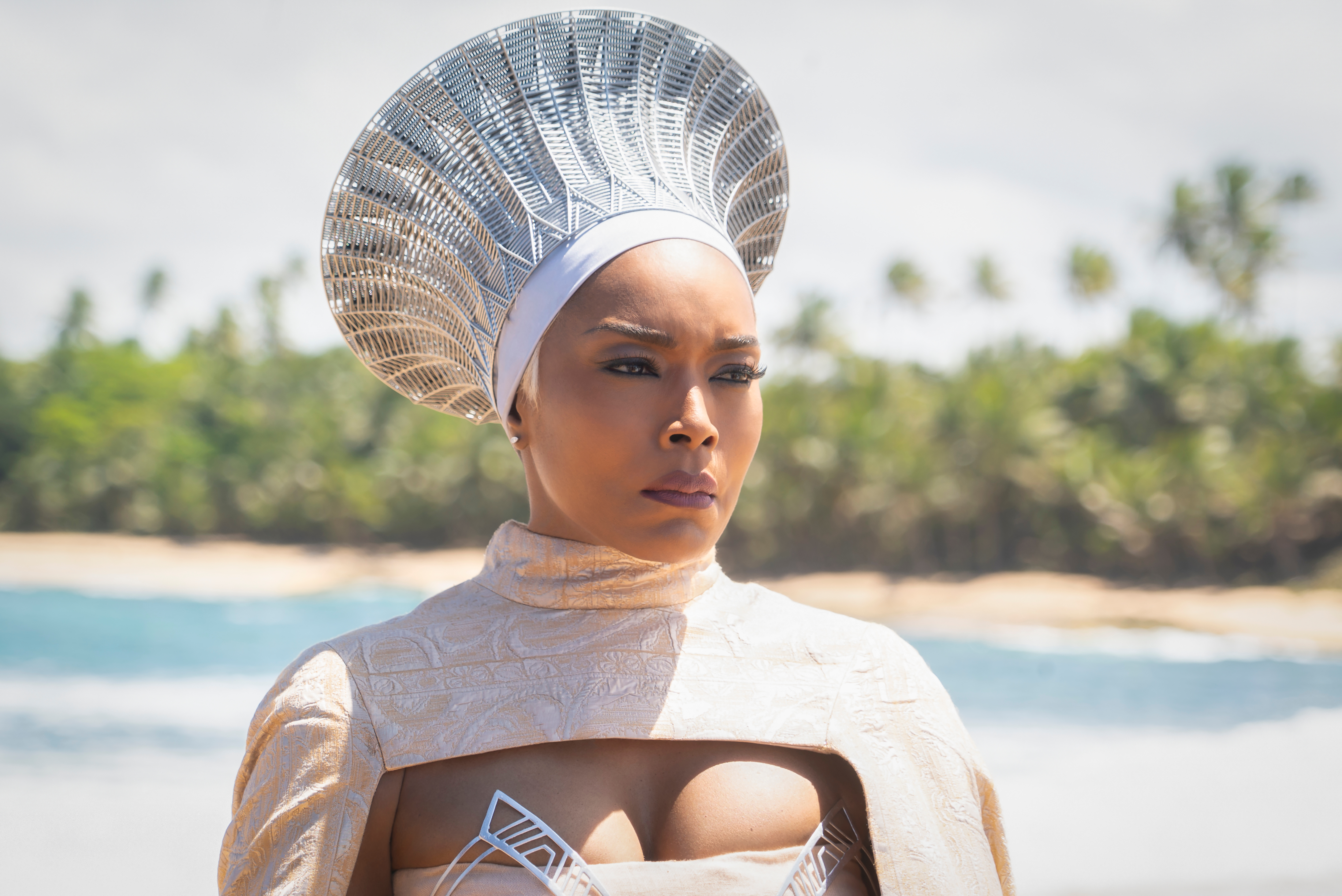 A woman wearing an elaborate headdress and a white costume stands on a beach with a body of water in the background.