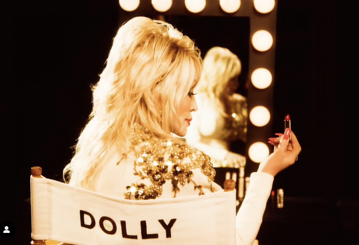 A woman with long blonde hair wearing a gold sequined dress and applying red lipstick in front of a mirror with lights around the frame.