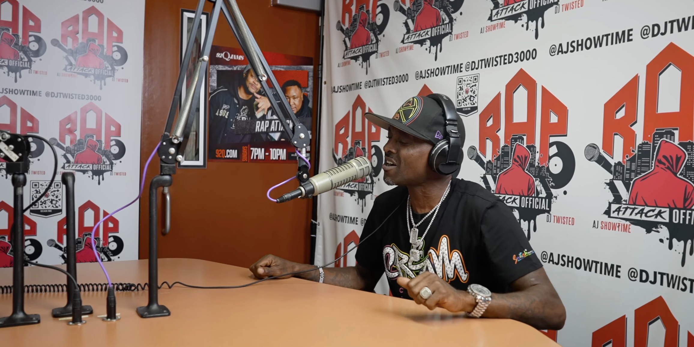 A person wearing headphones and a hat speaking into a microphone in a studio setting, with "Rap Attack" branding visible on the walls.